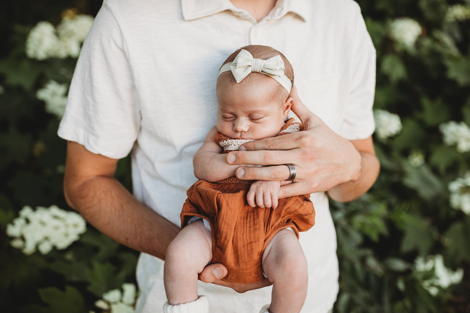 An infant baby girl sleeps in dad's hands against his chest while standing in a garden