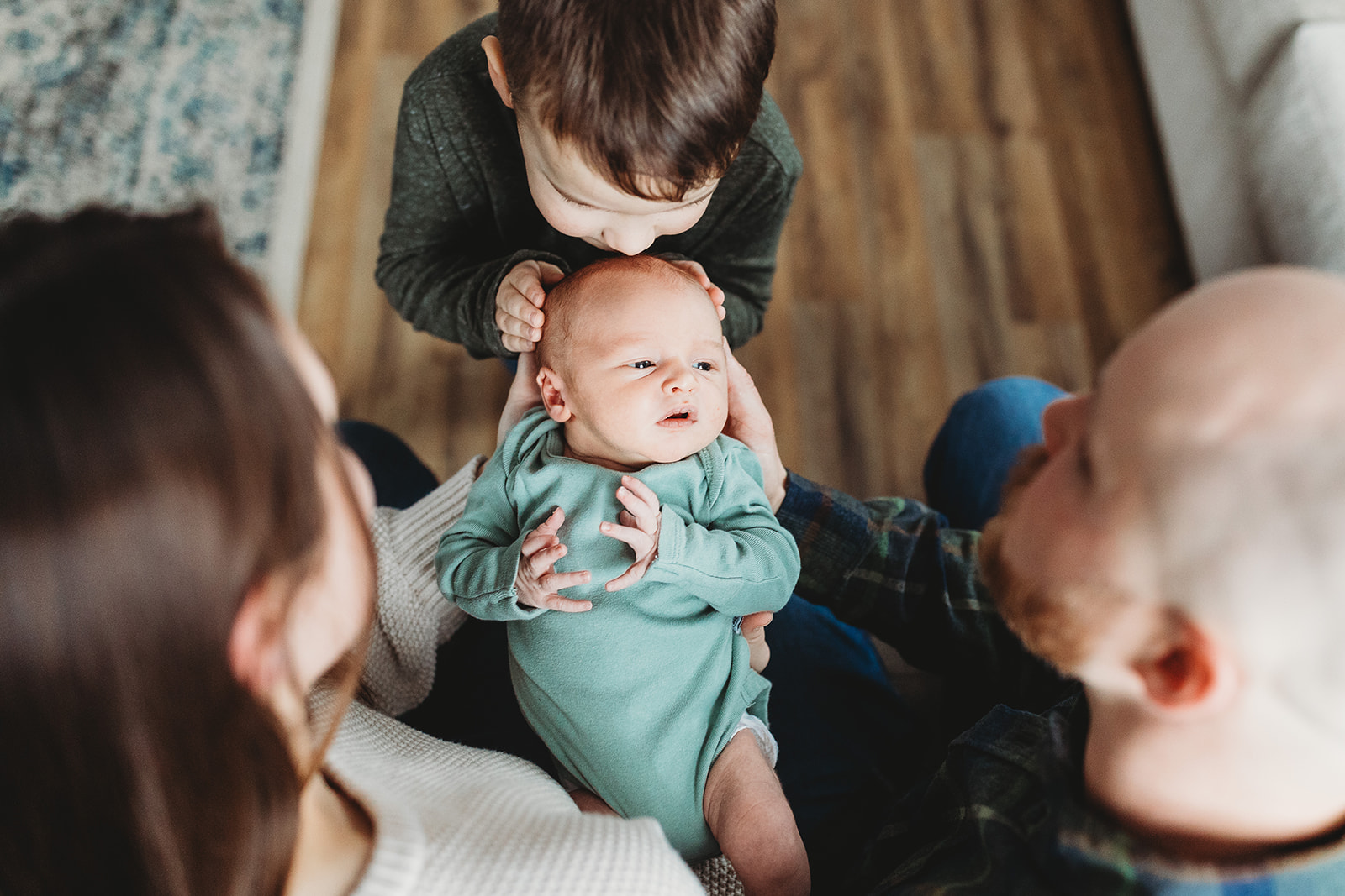 A toddler boy in a green sweater kisses the head of his newborn baby sibling in mom and dad's lap after leaving the Birthing Centers Harrisonburg, VA