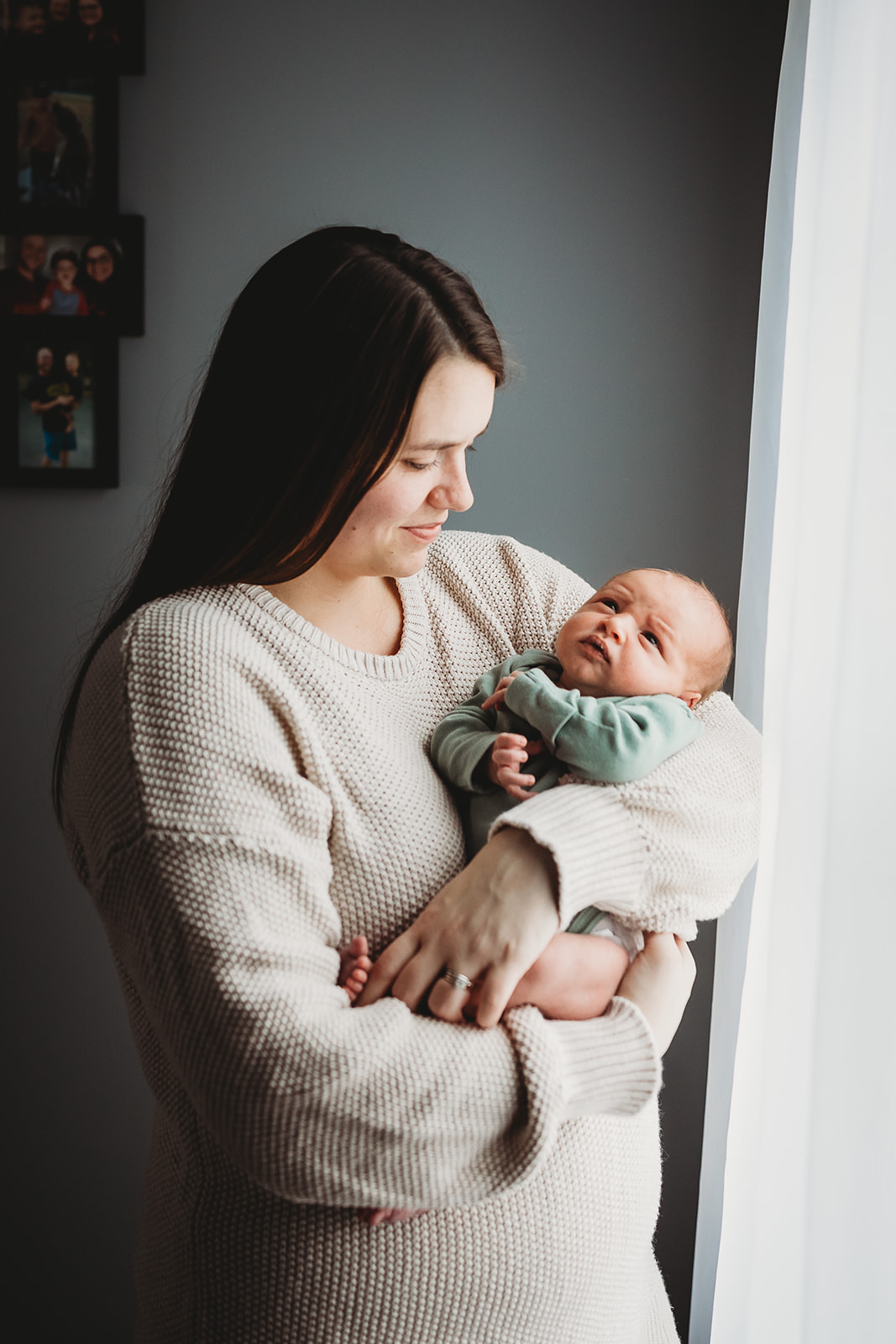 A happy mom in a white sweater stands in a window cradling her newborn baby in her arms thanks to Birthing Centers Harrisonburg, VA