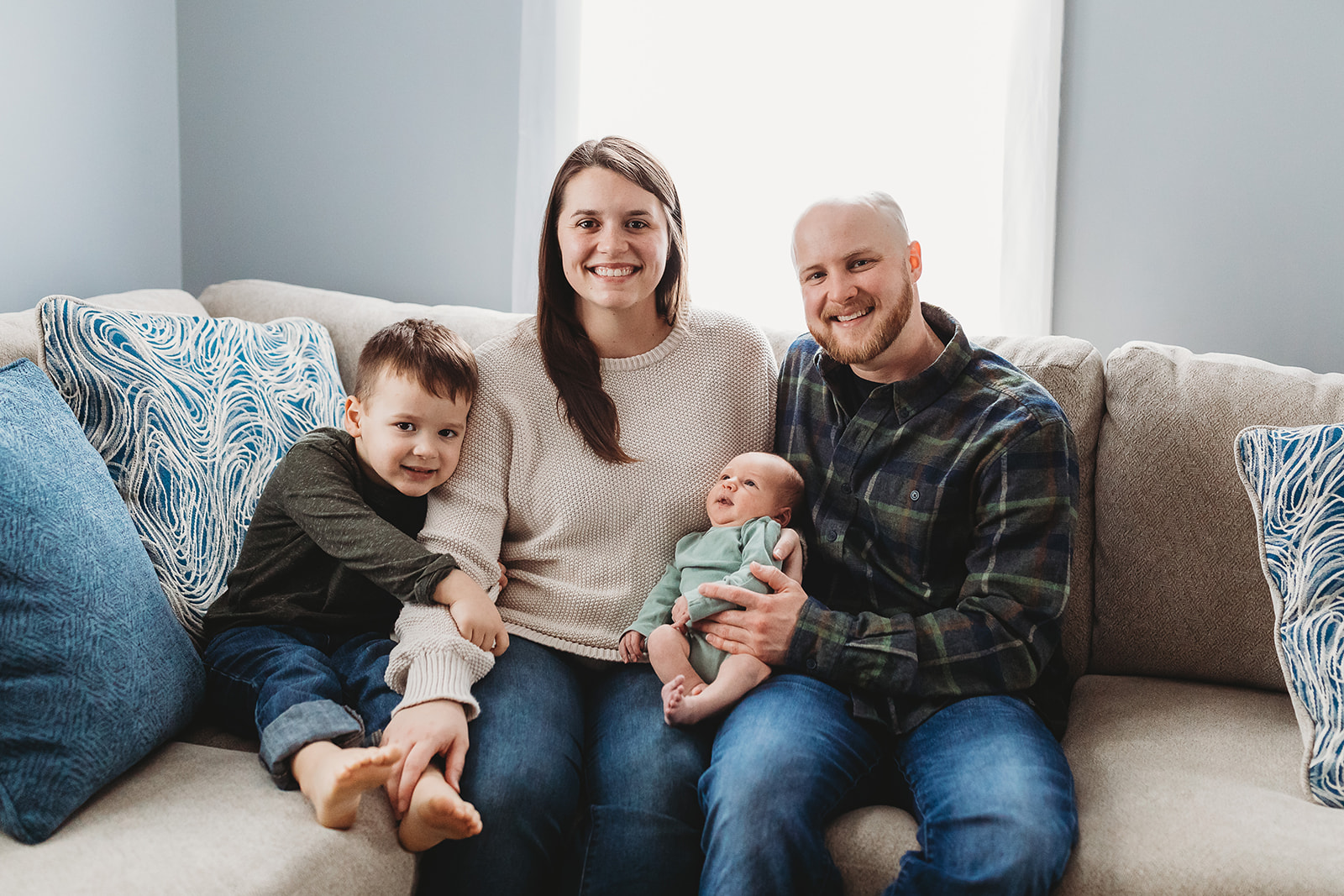 A happy family sits on a couch with a toddler and newborn baby