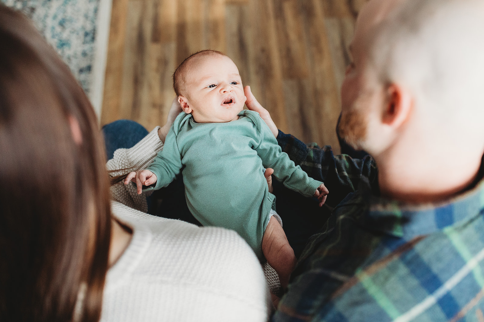 A newborn baby looks up at dad while laying in mom and dad's laps on a couch