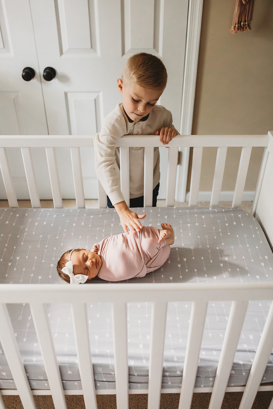 A toddler boy reaches in to touch his sleeping newborn baby sister in a tight pink swaddle