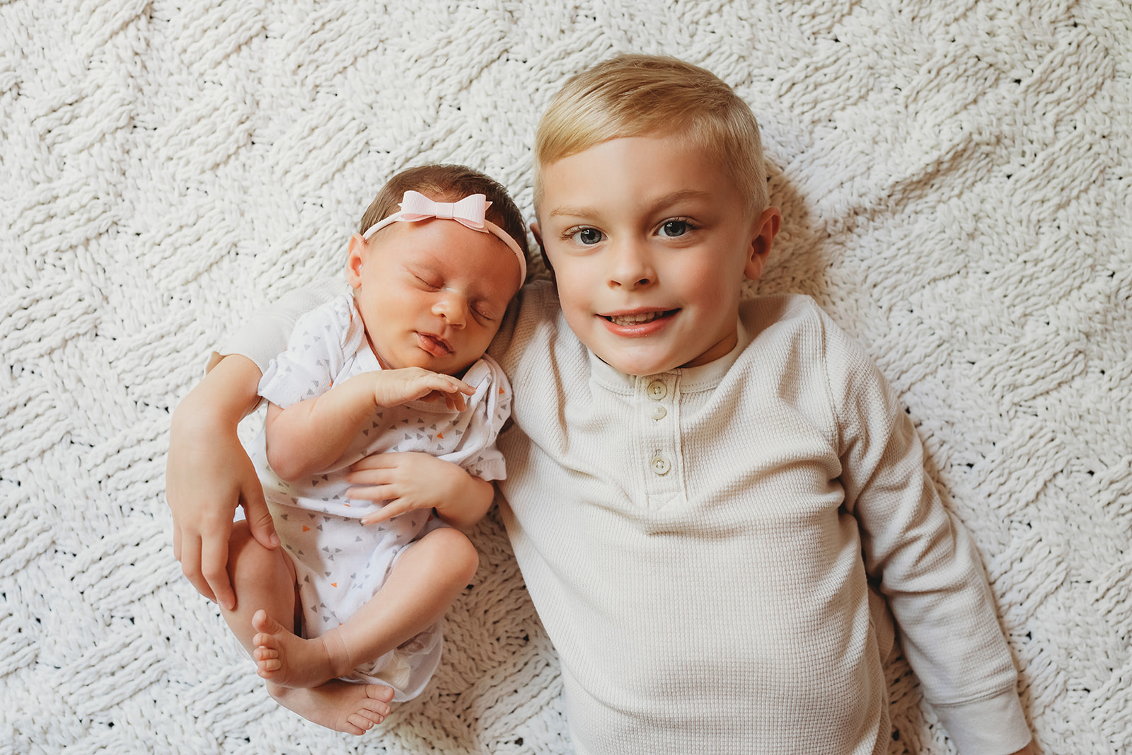 A toddler boy lays on a bed with one arm aroud his newborn baby sister