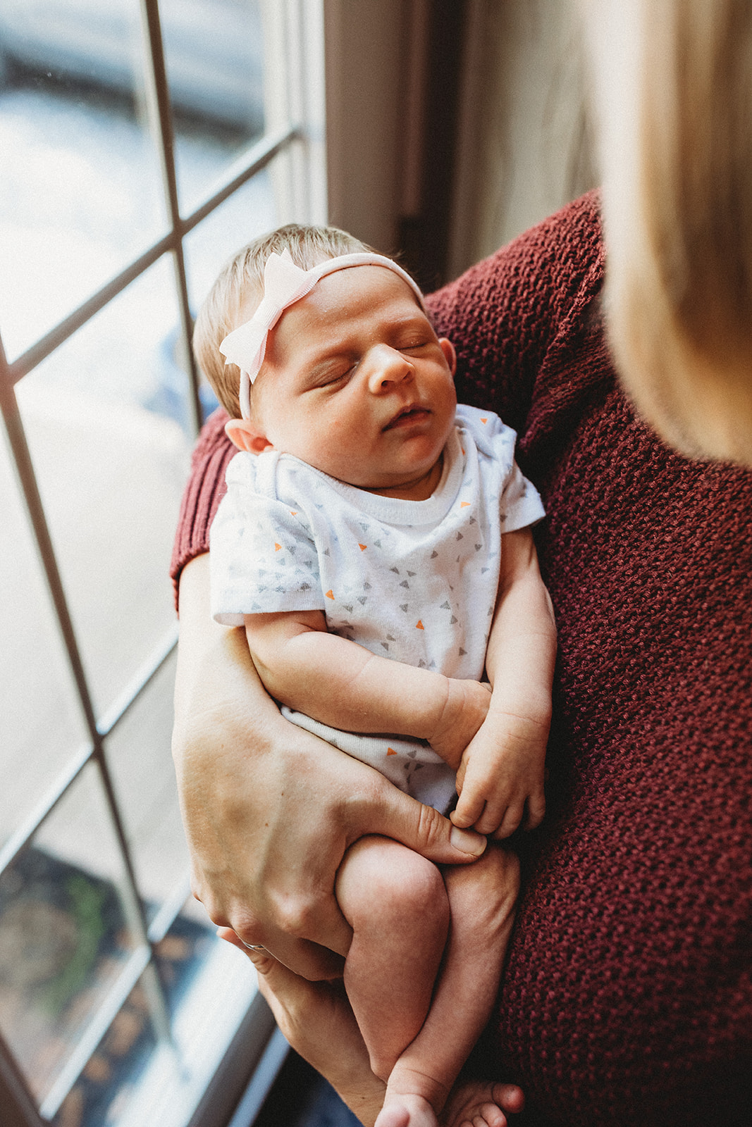 A newborn baby girl sleeps in mom's arms while she stands in a window in a red sweater after leaving Brookhaven Birth Center