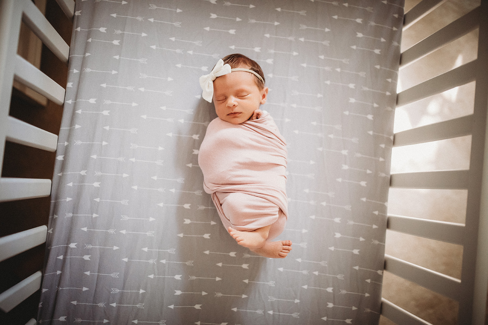 A newborn baby girl in a pink swaddle and white bow headband sleeps in a crib with arrow sheets after using Brookhaven Birth Center