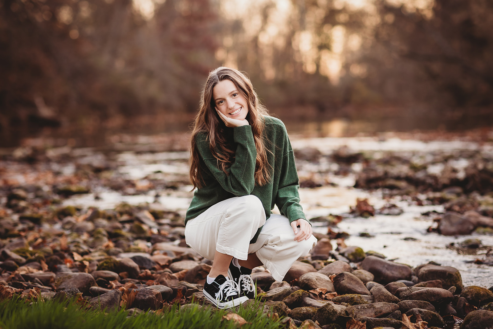 A high school senior squats on a rocky riverbed at sunset in a green sweater and white pants after visiting Clothing Stores in Harrisonburg, VA