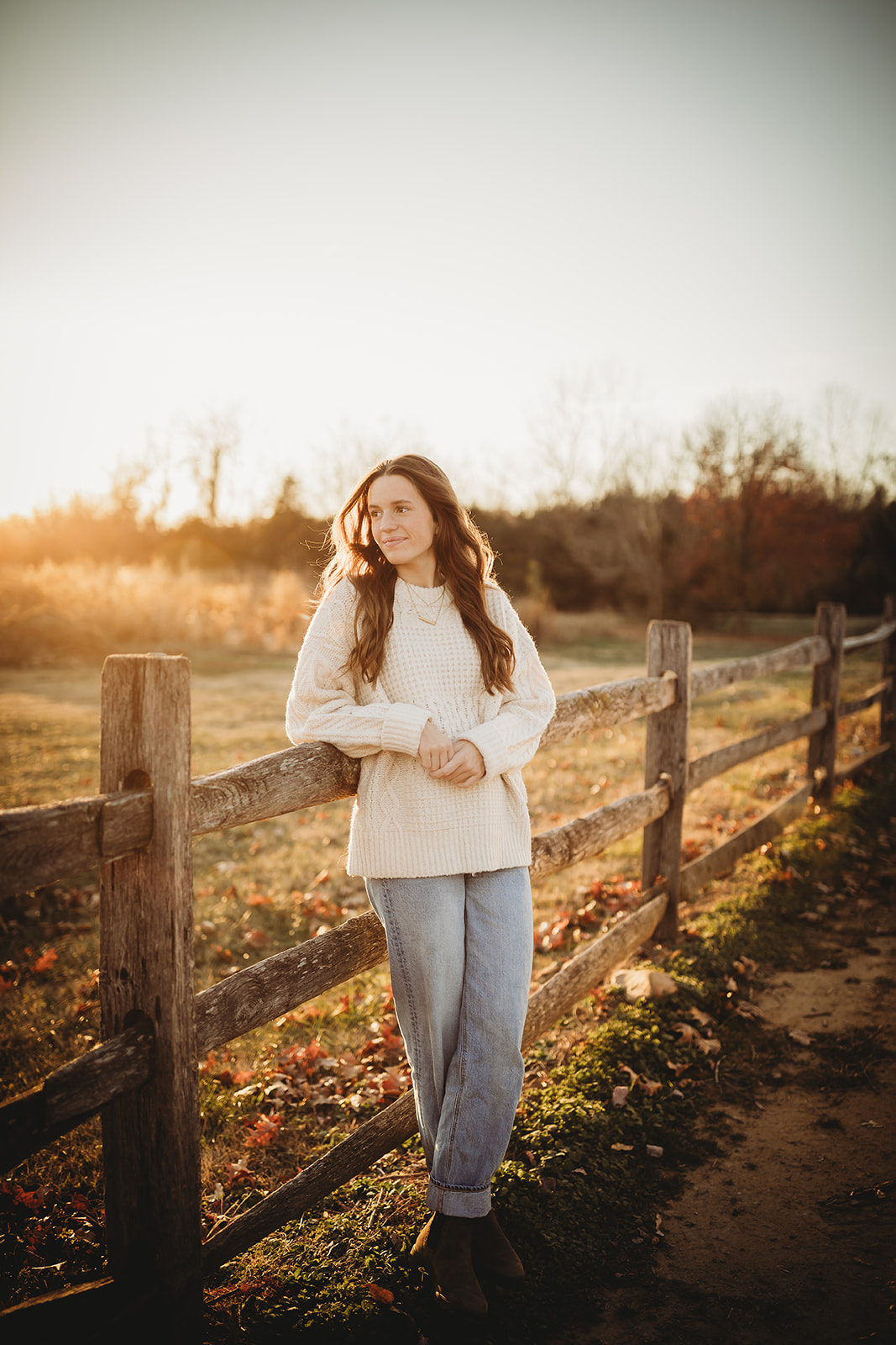 A high schools senior in a white sweater leans on a wooden farm fence at sunset after visiting Clothing Stores in Harrisonburg, VA