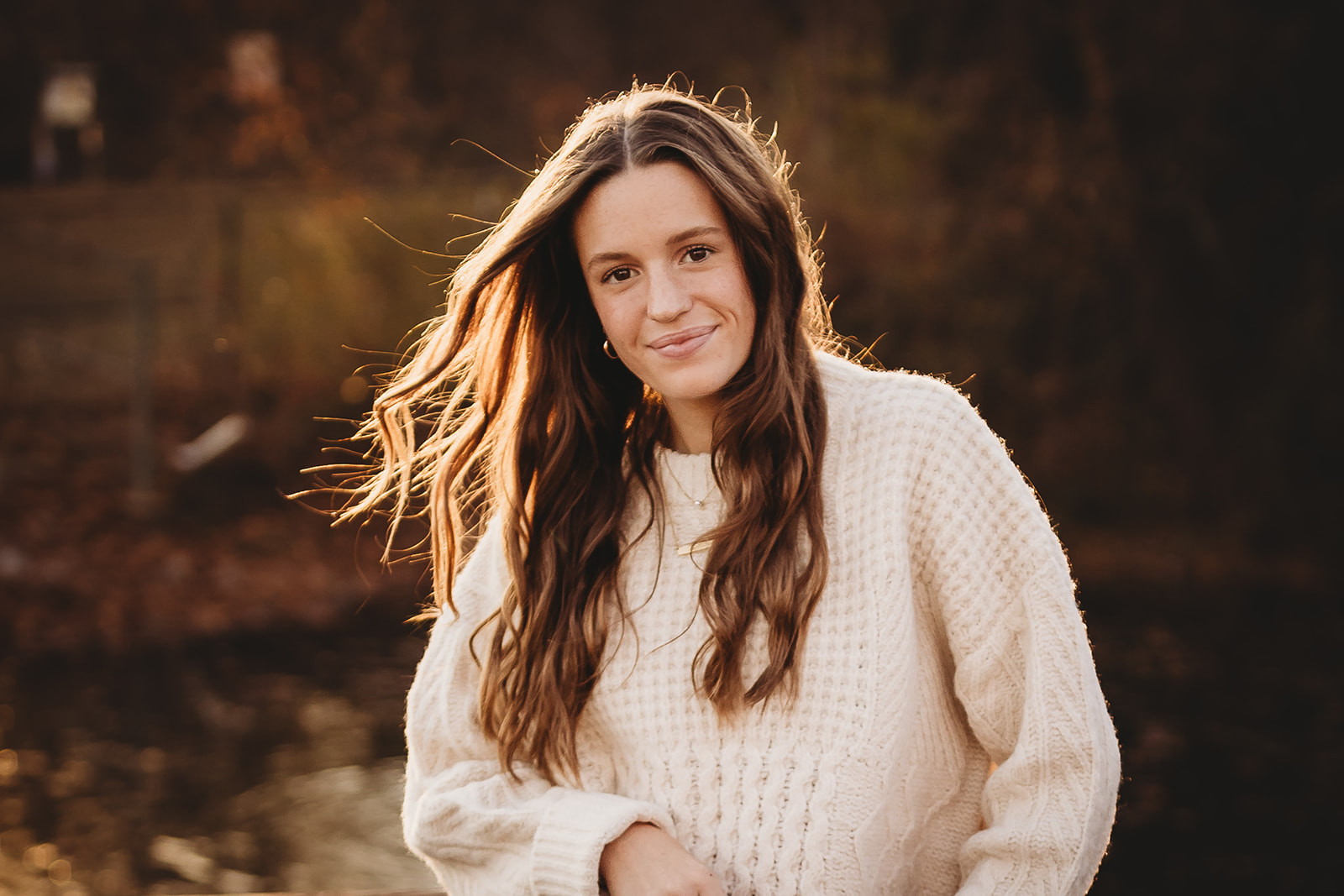 A woman stands against a windy fence in a white knit sweater at sunset