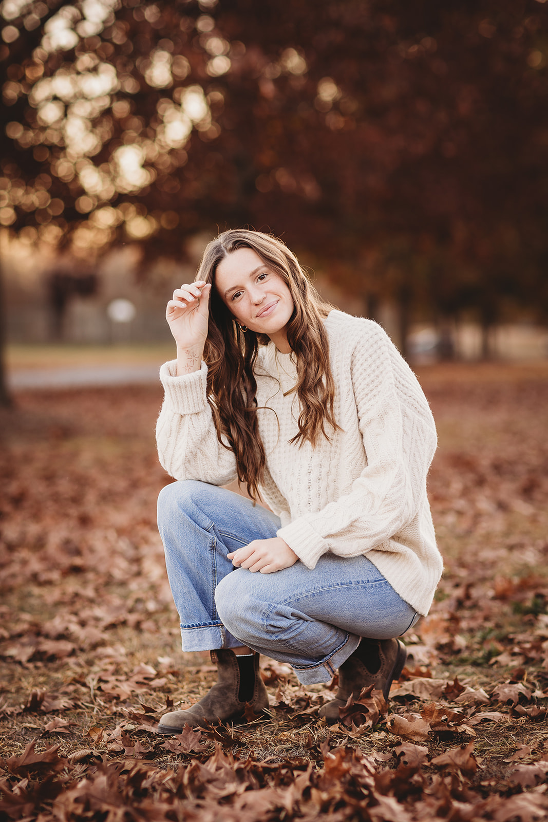 A high school senior in a white sweater kneels in some fallen leaves at sunset in a park