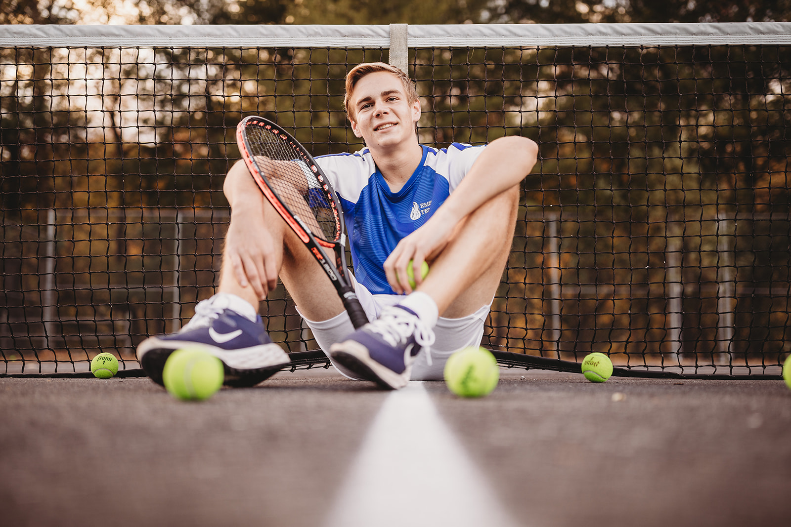 A high school senior sits on a tennis court with balls and a racket after exploring Colleges Near Harrisonburg, VA