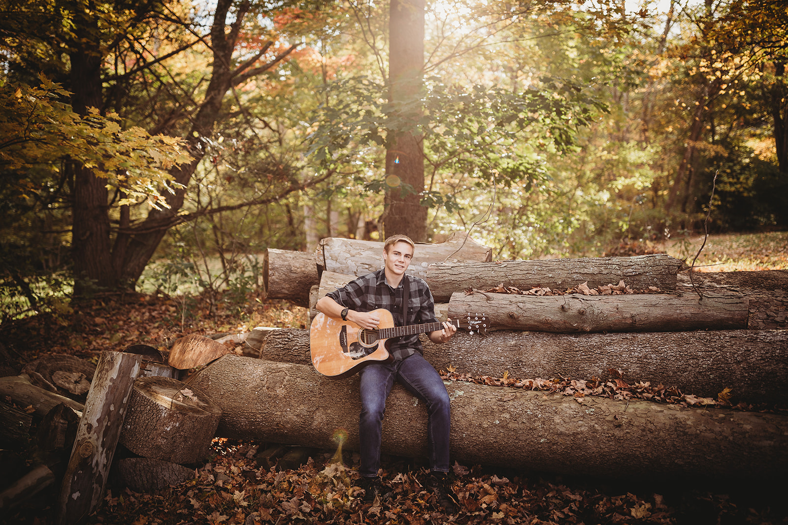 A teenage boy sits on a fallen log playing guitar in a forest at sunset