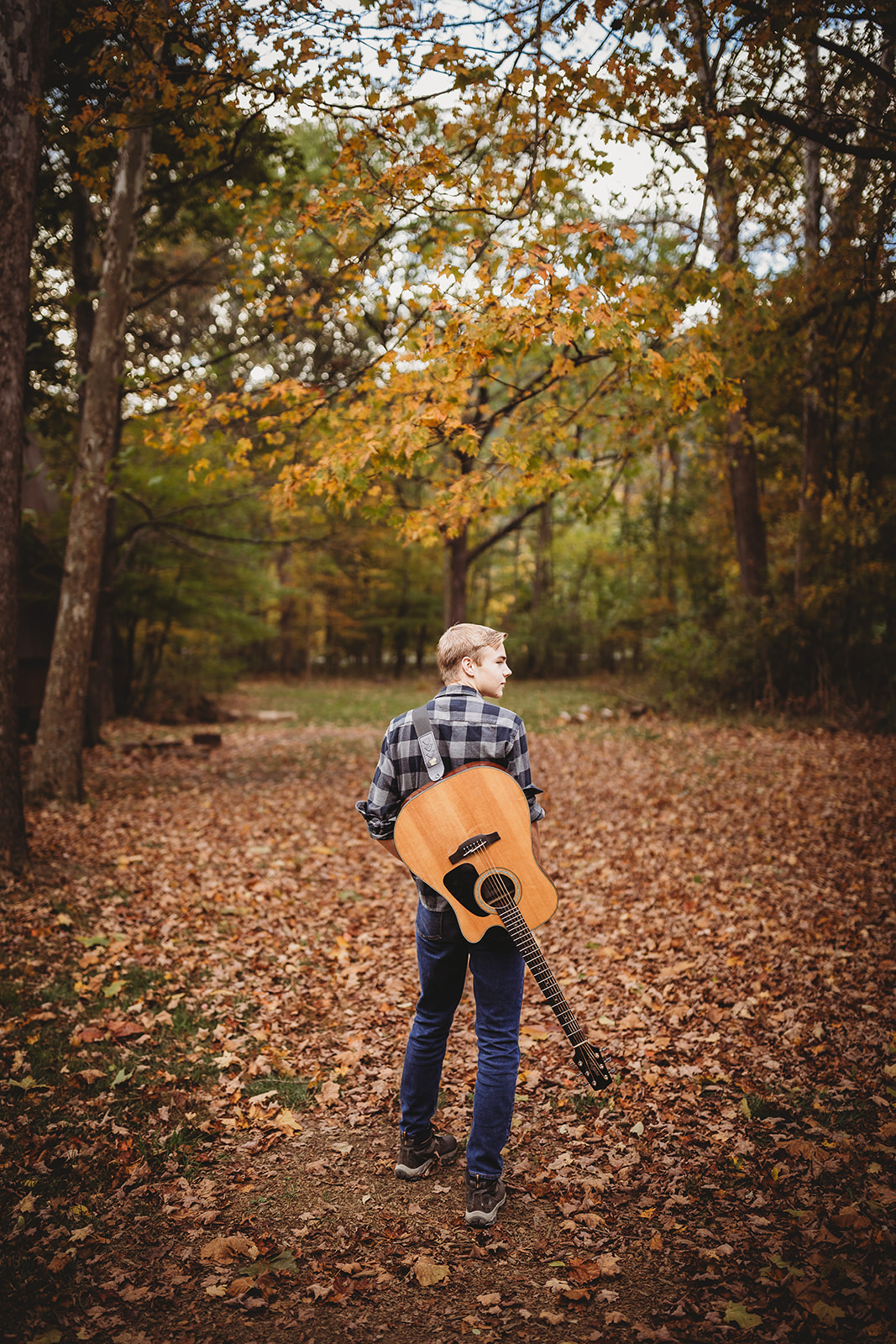 A guitar player in jeans walks in a leaf covered trail with guitar on his back