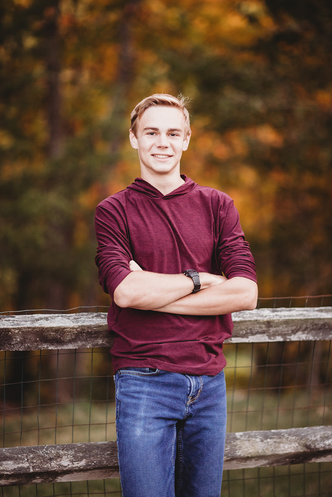 A high school senior in jeans and a maroon shirt leans on a farm fence at sunset with arms crossed after exploring Colleges Near Harrisonburg, VA