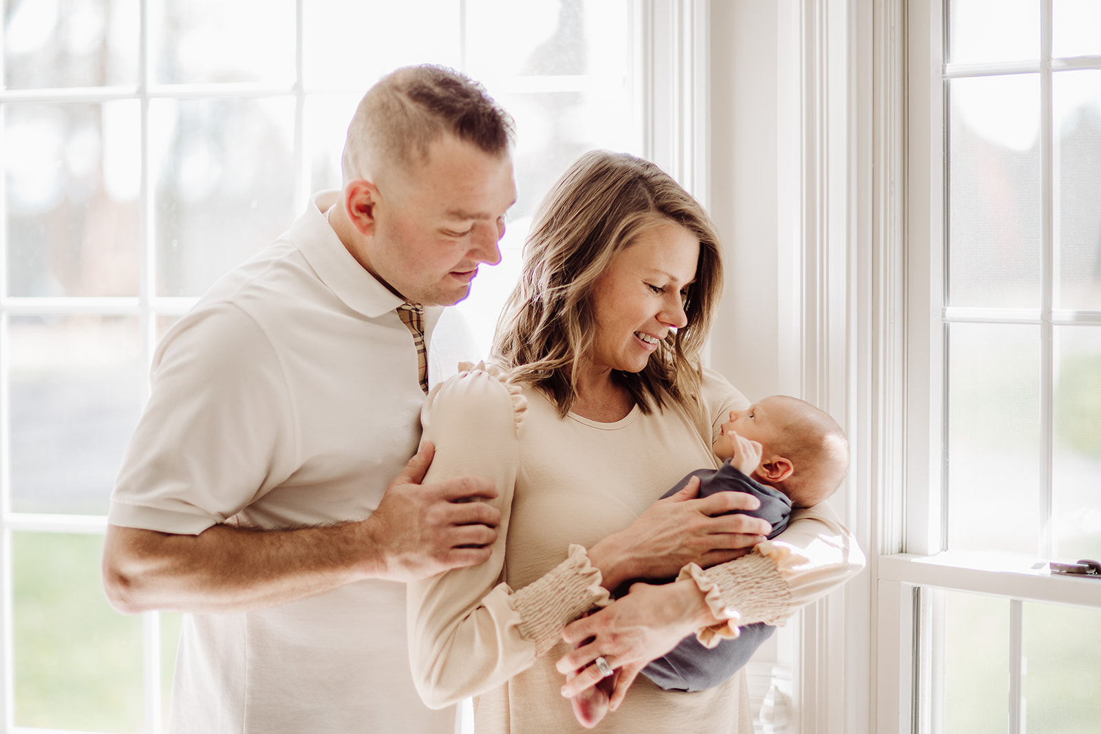 Happy new parents stand in a window smiling down at their awake newborn baby in mom's arms before visiting Daycares Harrisonburg VA