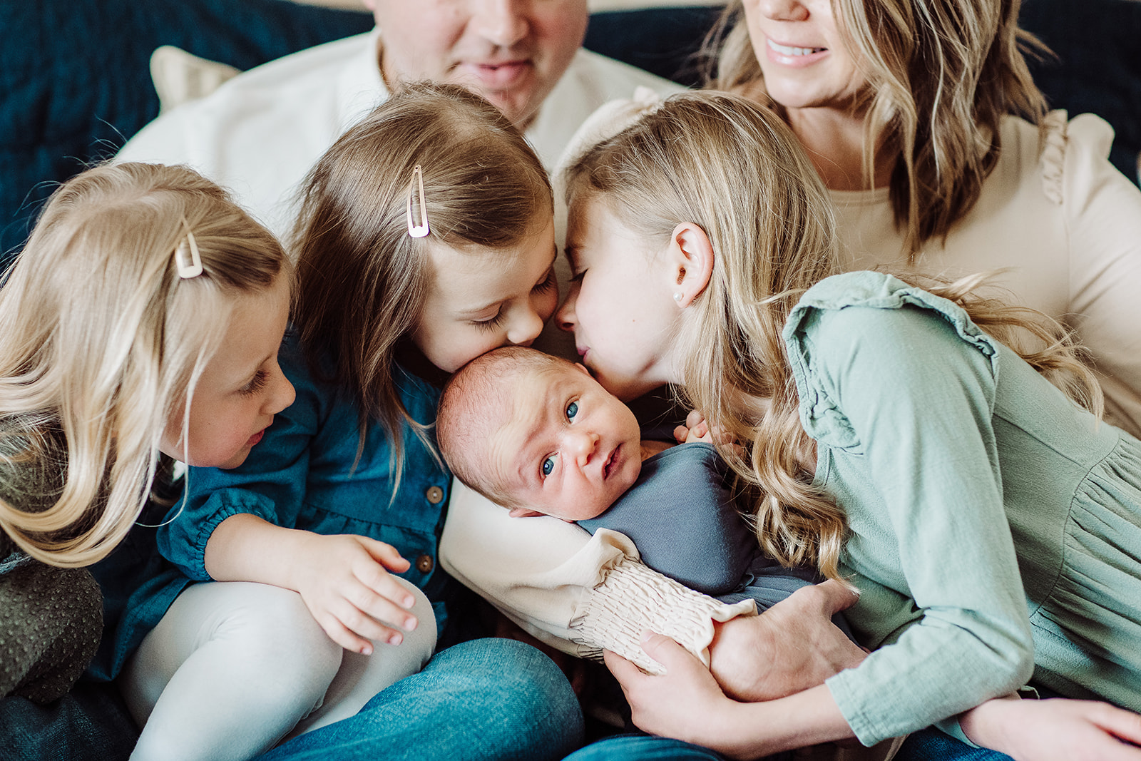 Three toddler sisters kiss their newborn baby sibling in mom and dad's lap