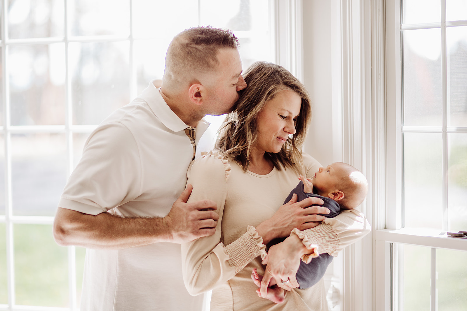 A father kisses his wife's head as she cradles their newborn baby in a window