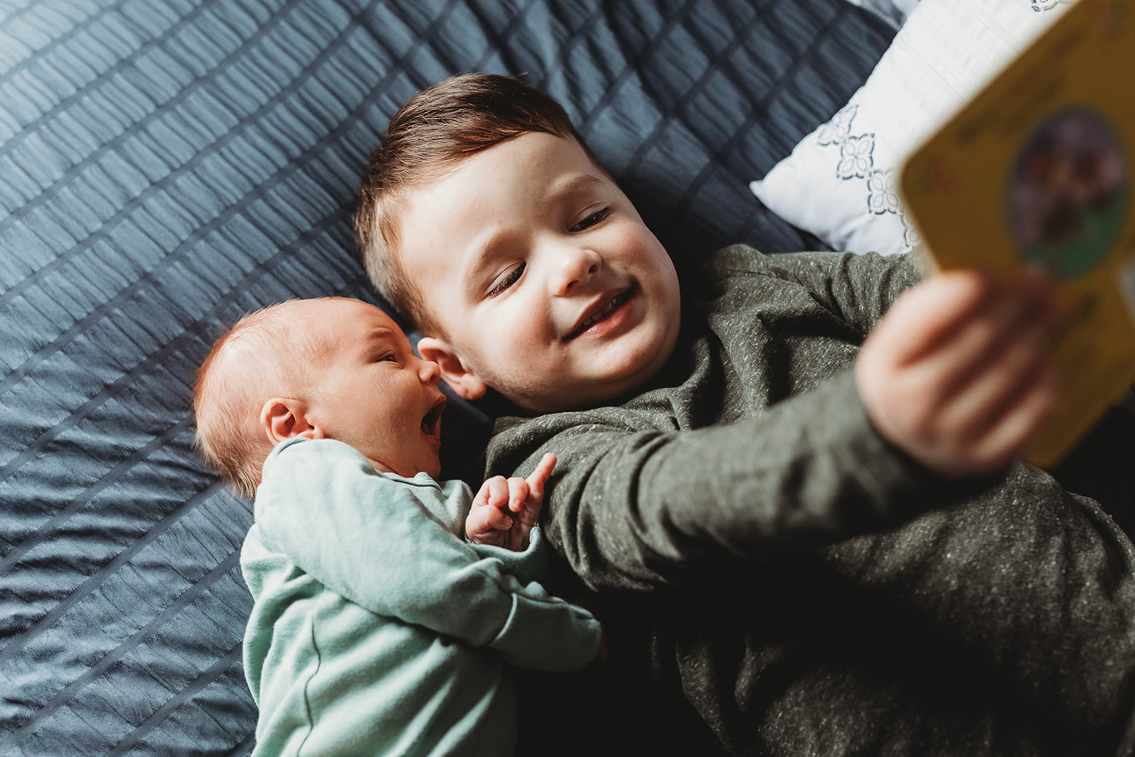 A toddler boy reads a book to his newborn sibling while laying on a bed after using Genesis Midwifery