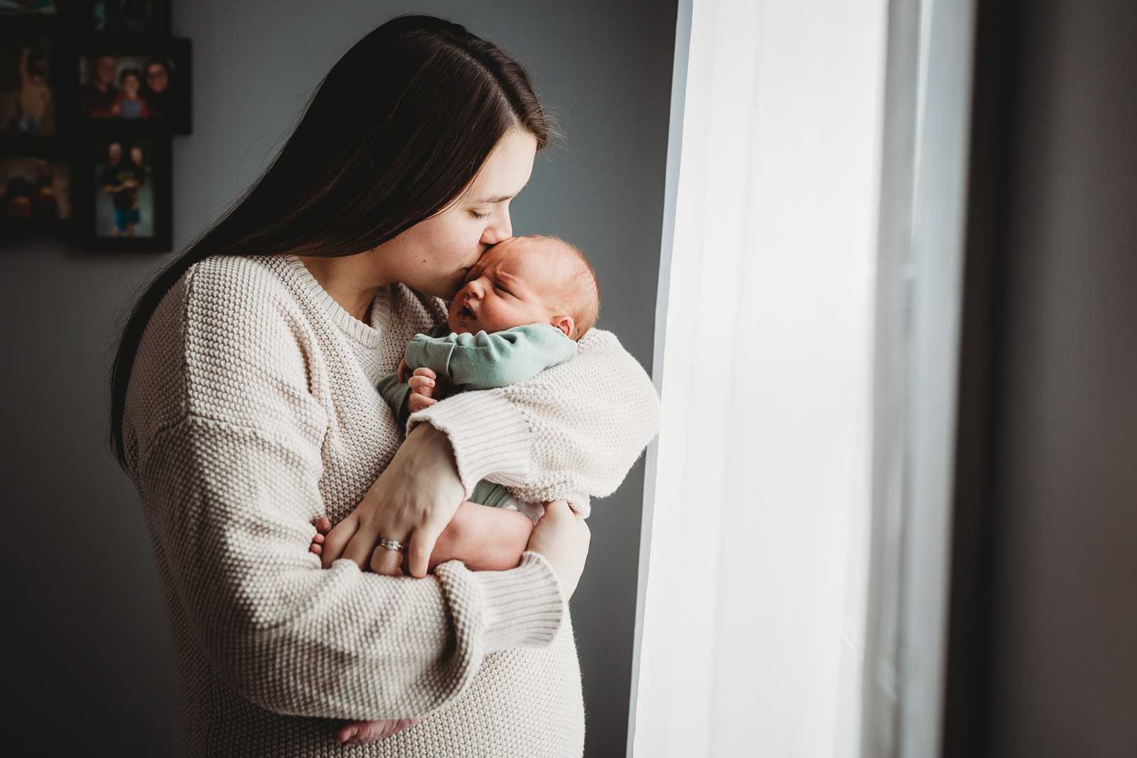 A new mom in a tan sweater kisses the head of her newborn baby while standing in a window