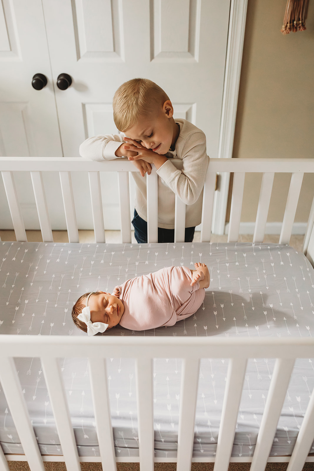 A toddler boy leans on a crib looking down on his sleeping newborn baby sister