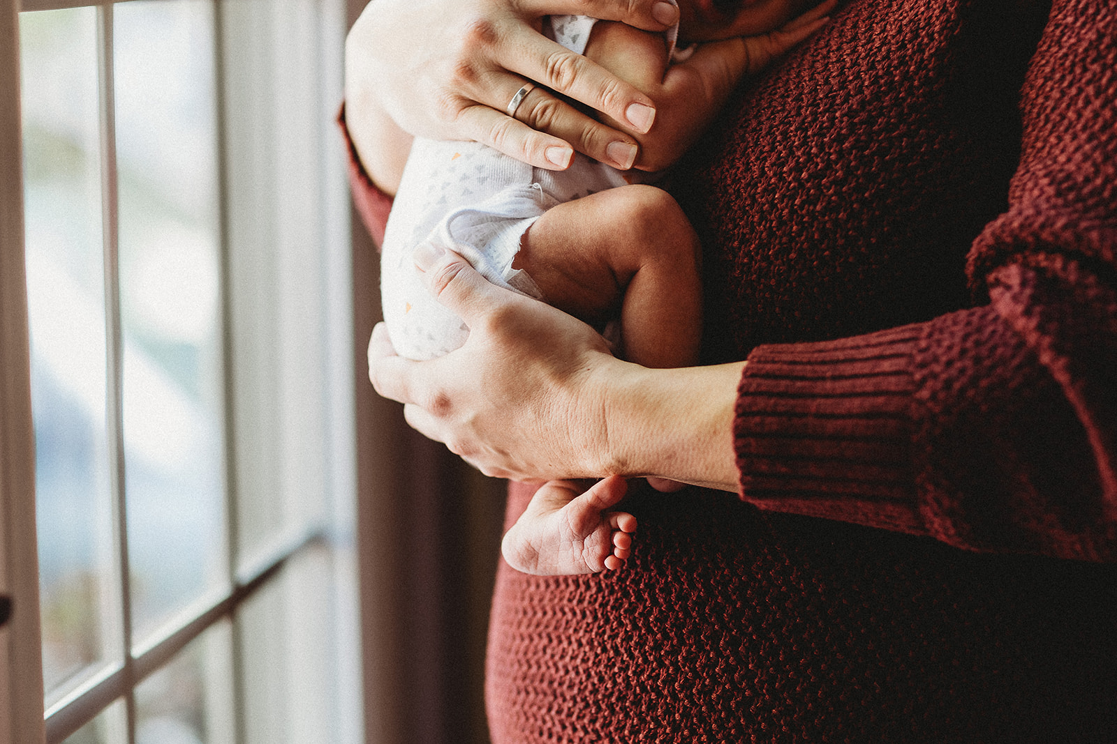 Details of a mother in a red sweater standing in a window holding her newborn baby thanks to Midwives Harrisonburg, VA