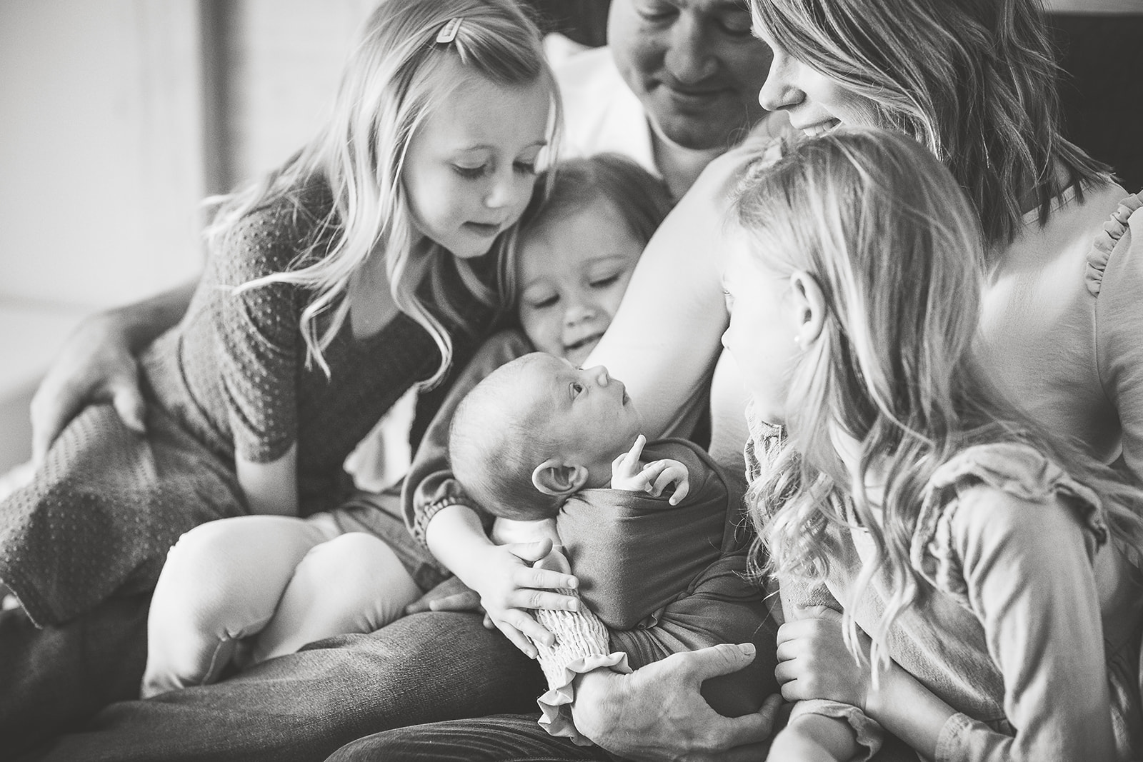 A newborn baby looks up at mom from her lap while sitting on a bed with dad and three toddler sisters