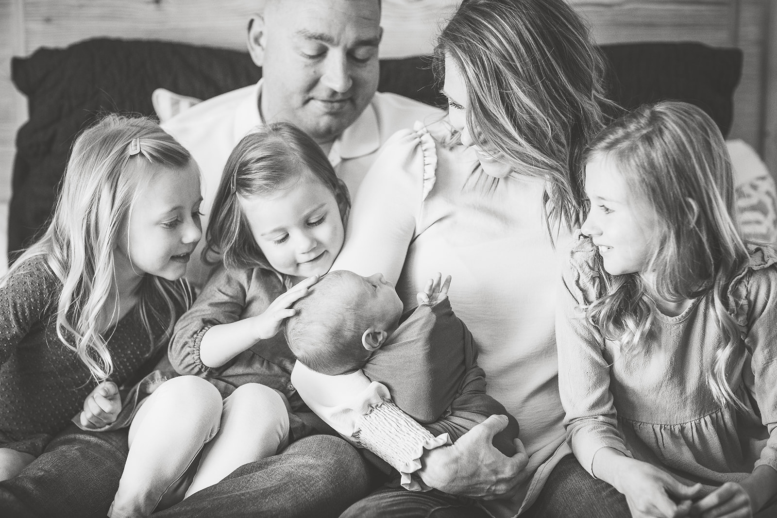 Three toddler sisters sit in mom and dad's laps on a bed while playing with their newborn baby sibling