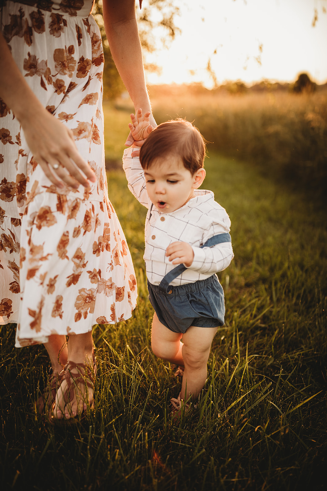 A toddler boy explores a grassy trail at sunset while holding mom's hand