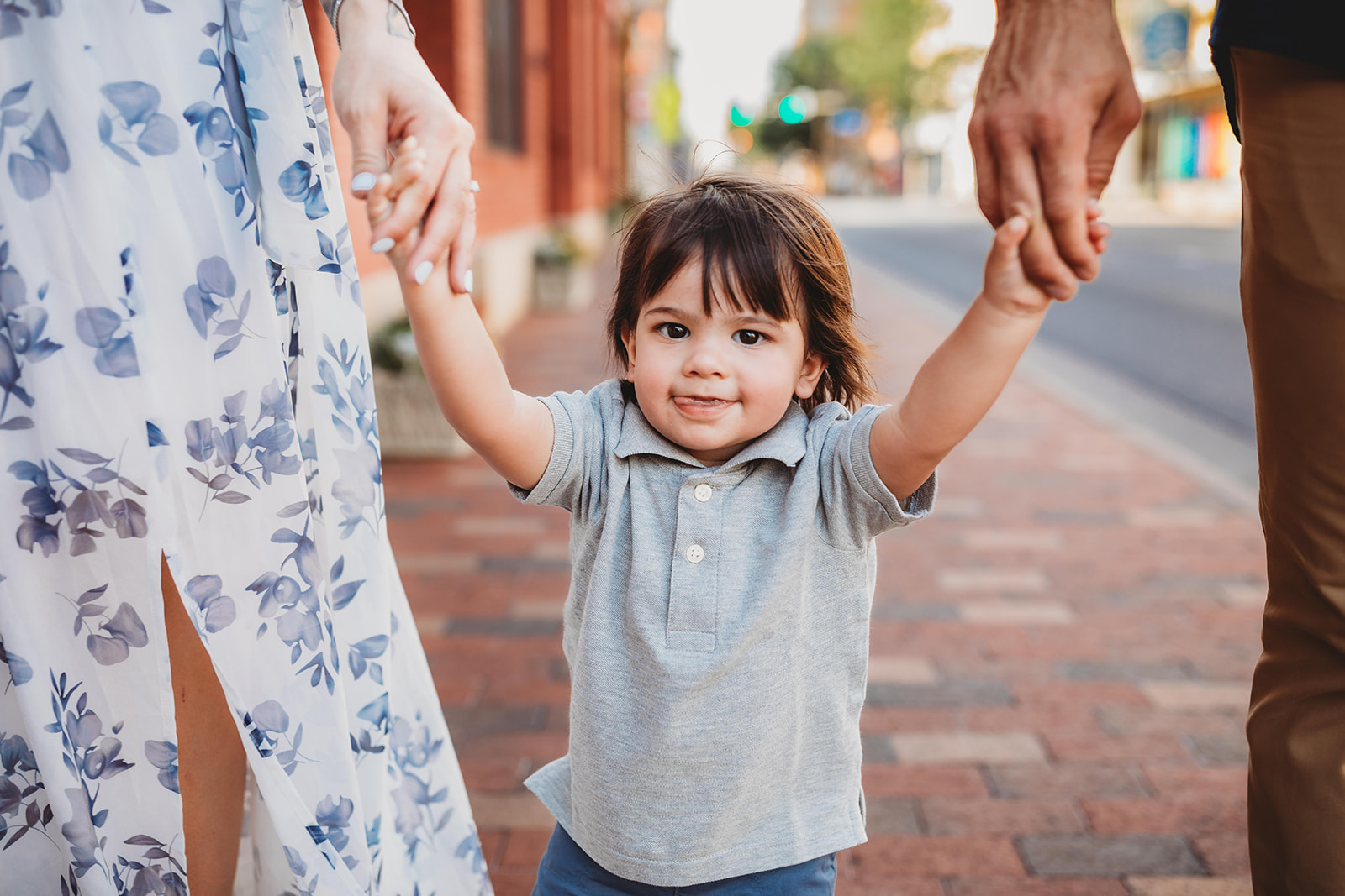 A toddler boy with dark hair and a grey polo walks down a brick sidewalk holding mom and dad's hands before visiting a Pediatric Dentist Harrisonburg, VA