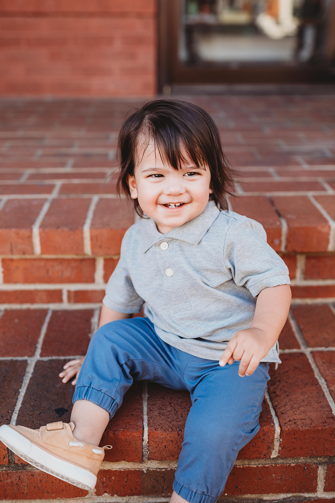 A toddler boy in a grey polo sits on brick steps smiling before visiting Pediatric Dentist Harrisonburg, VA