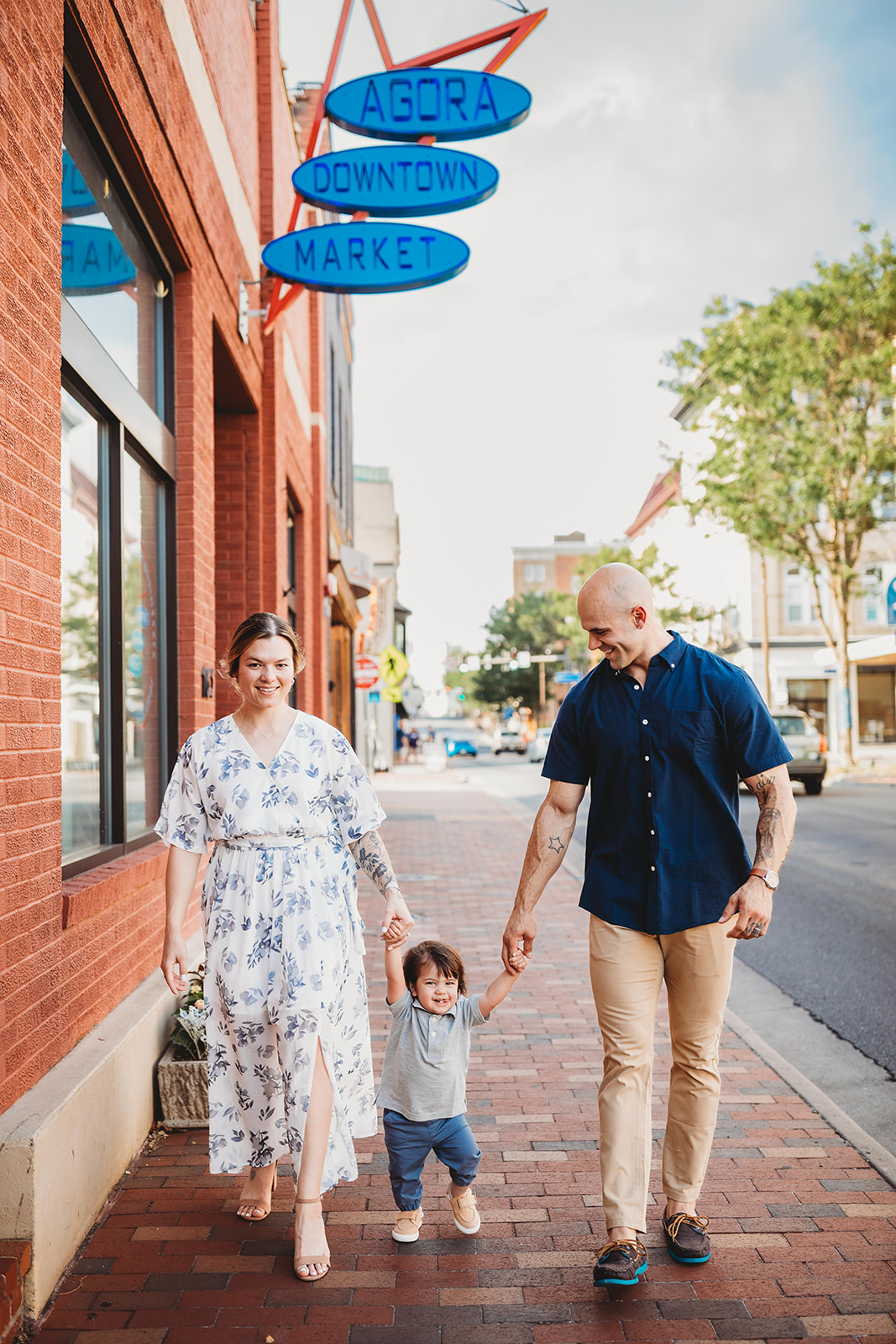 A mom and dad walk on a downtown sidewalk holding hands with their laughing toddler son after visiting a Pediatric Dentist Harrisonburg, VA