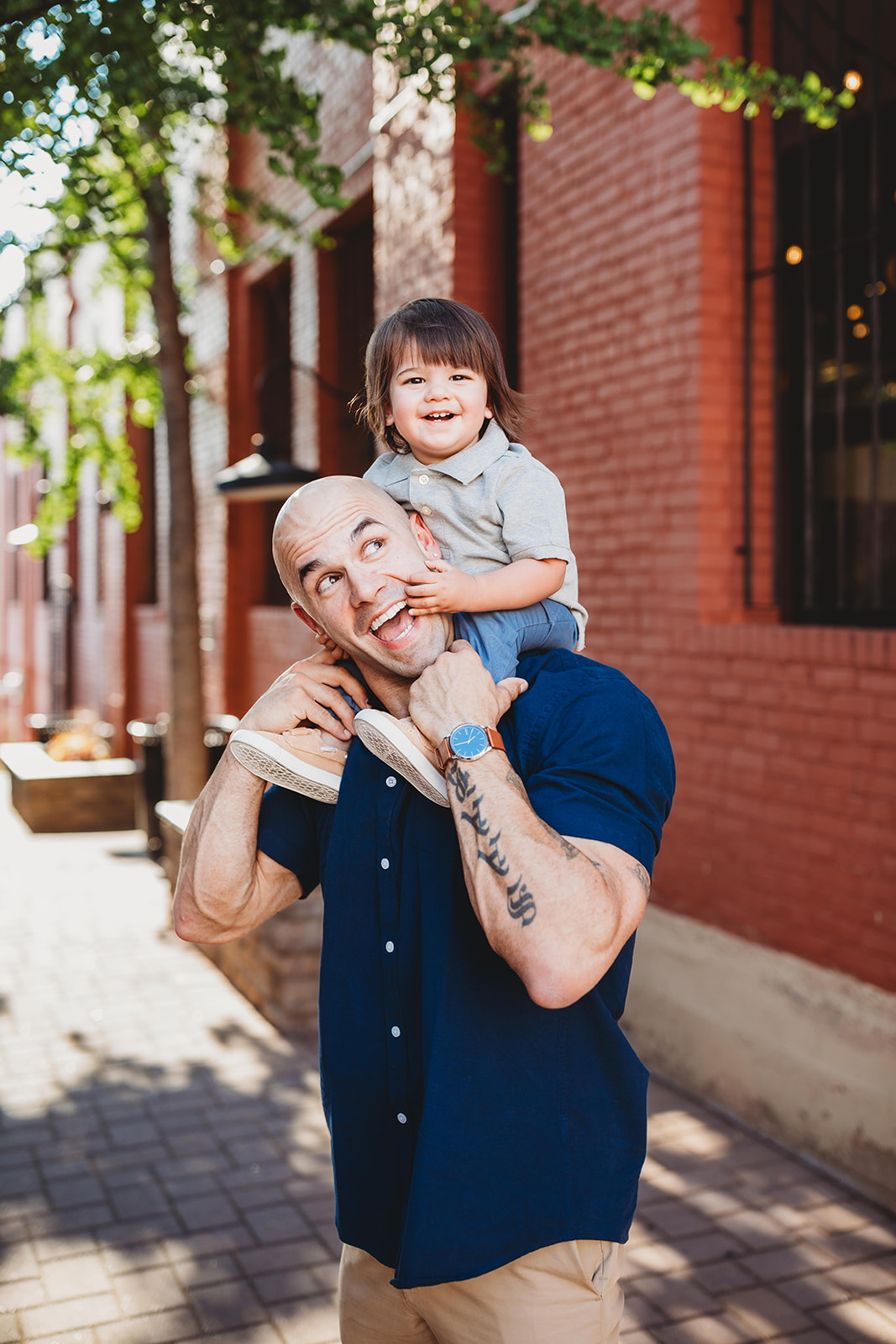 A laughing toddler boy sits on dad's shoulders in a downtown sidewalk