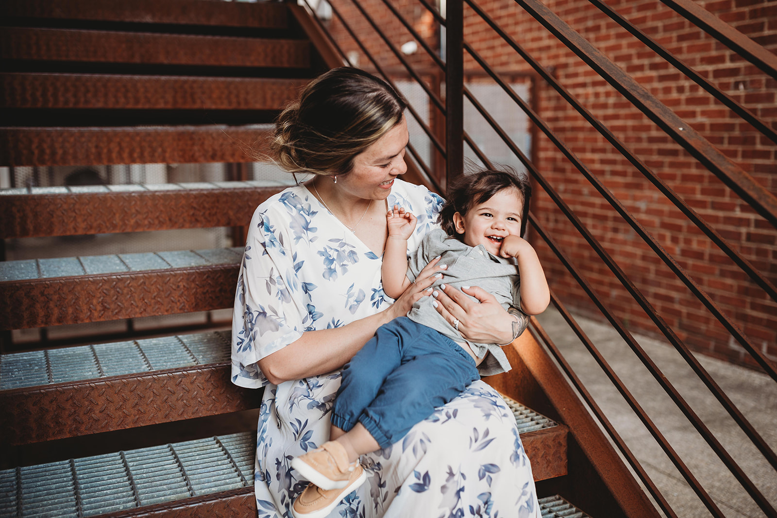 A mom sits on metal downtown stairs playing with her toddler son in her lap