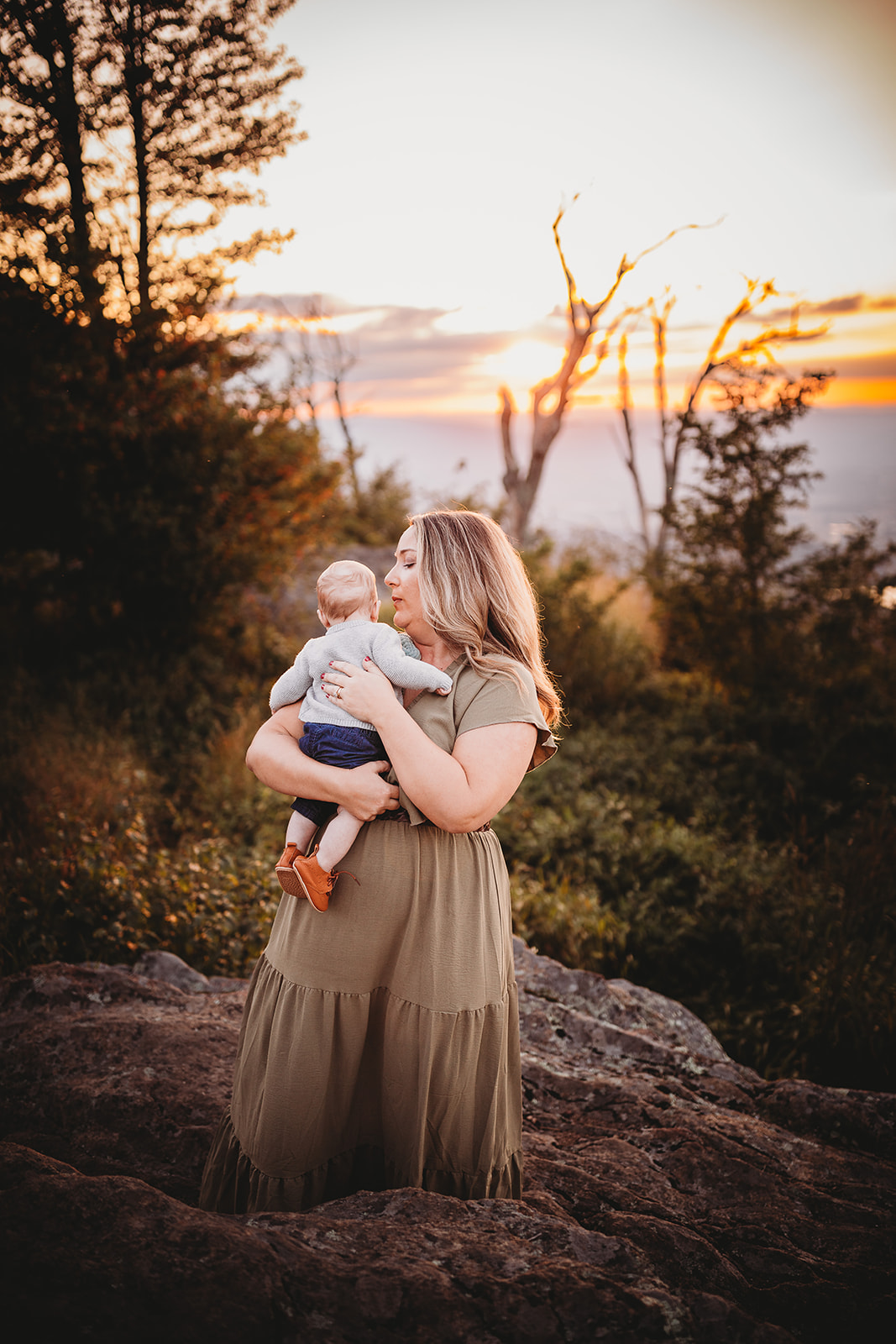 A mother ina. green dress stands on a mountain rock at sunset with her infant son on her shoulder after meeting Pediatricians Harrisonburg, VA