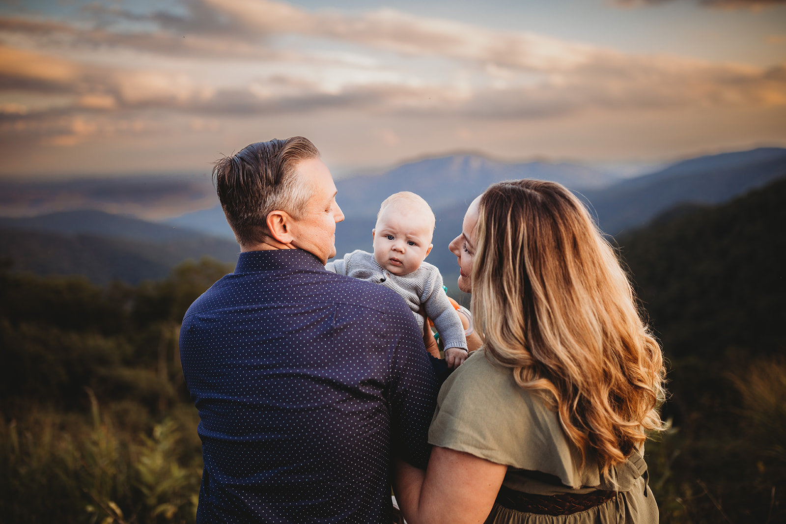 Happy parents stand on a mountain overlook with their infant between them