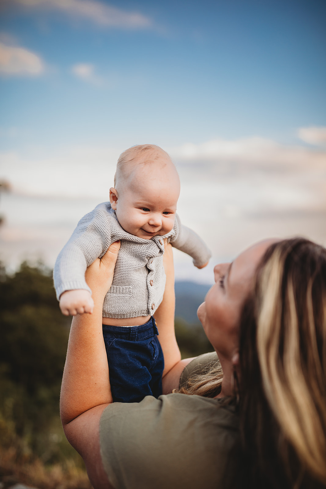 A giggling infant boy in a grey knit sweater is lifted by mom in the mountains at sunset