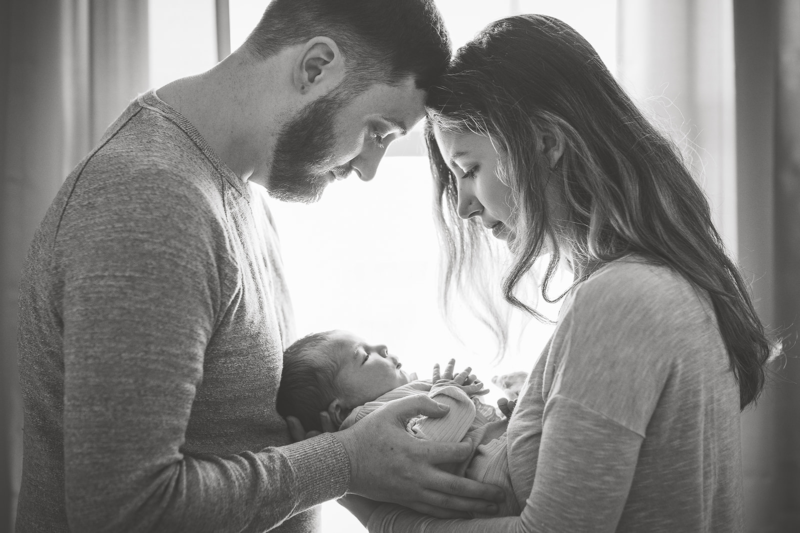 New parents touch foreheads and look down at their sleeping newborn baby between them