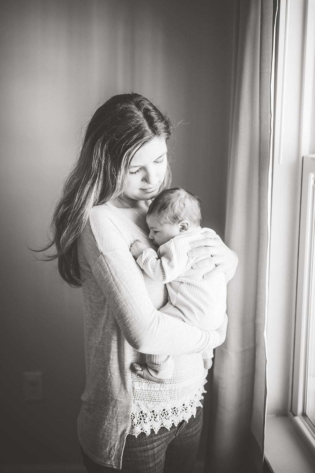 A new mother stands in a window cradling her newborn baby against her chest in black and white