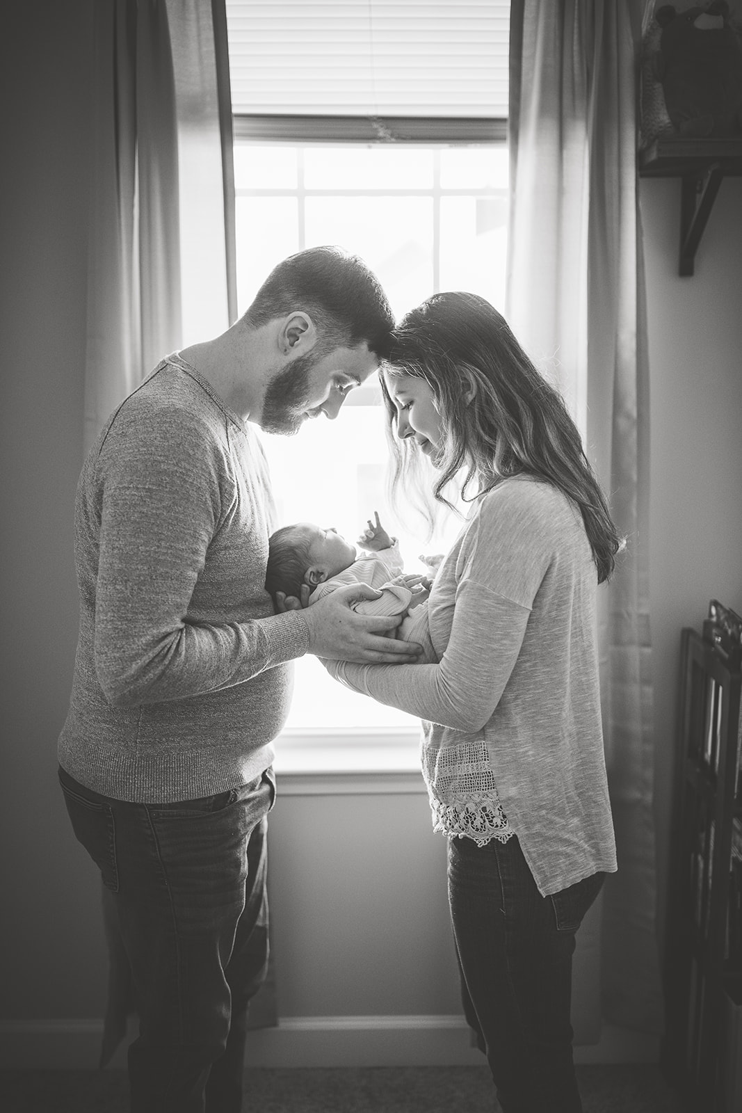 Happy new parents stand in a window holding their newborn baby between them after using Prenatal Massage Harrisonburg, VA