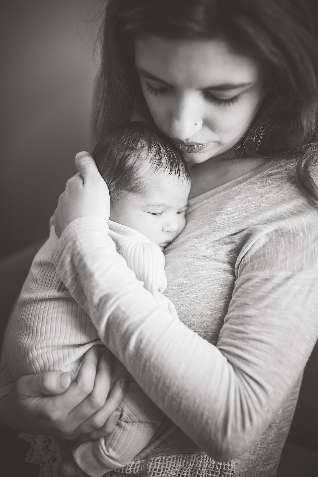 A new mother stands in a window cradling her newborn baby against her chest in black and white