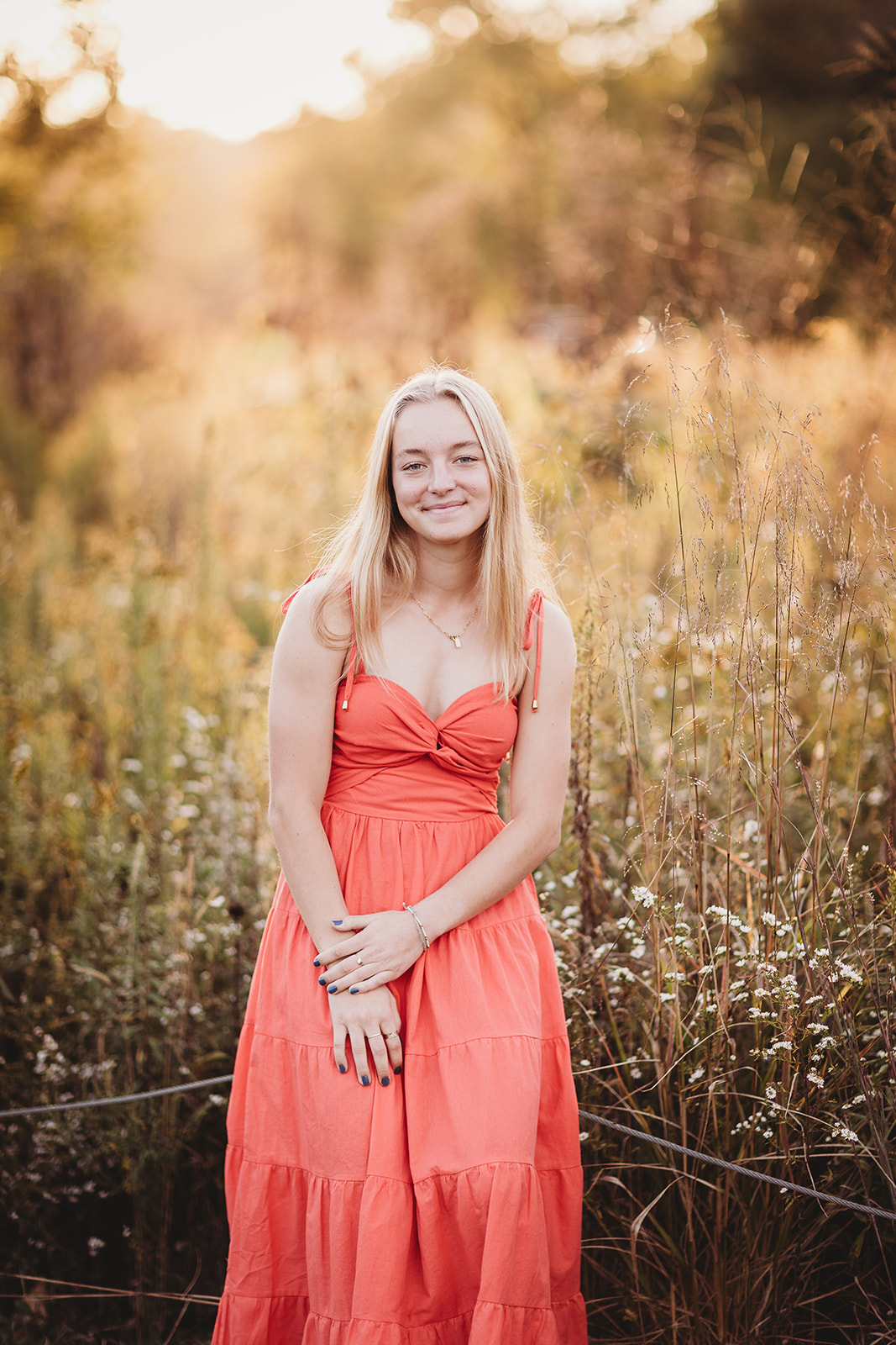 A teenage girl in a salmon dress stands by some tall golden grasses smiling before finding Prom Dresses Harrisonburg, VA