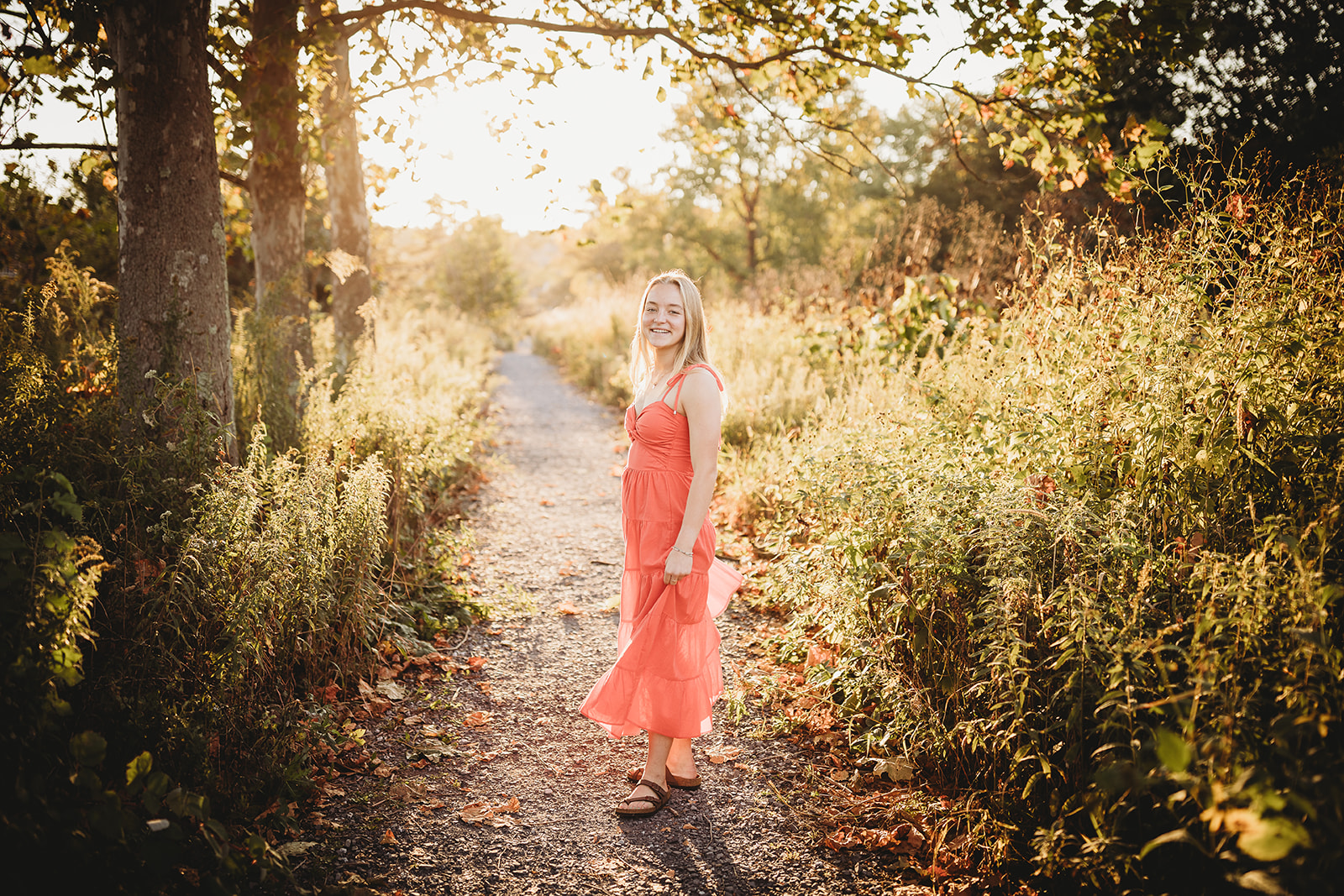 A high school senior turns and holds her dress while hiking in a trail at susnet