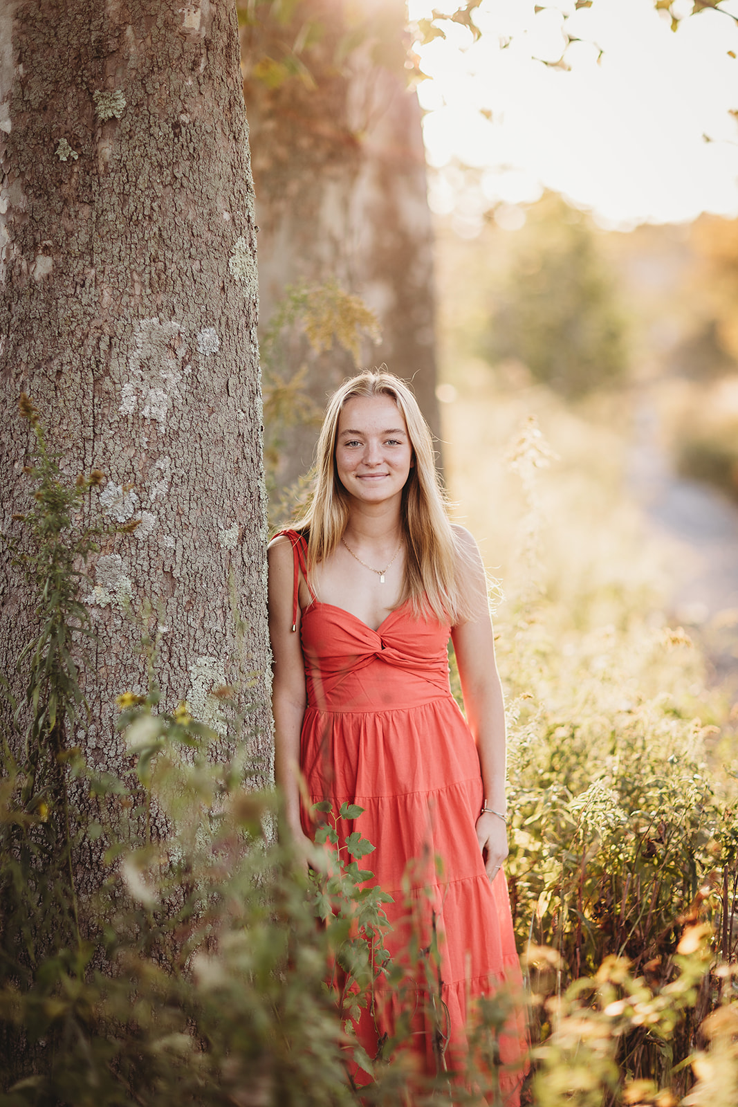 A high school senior leans against a large tree trunk at sunset smiling in a pink dress