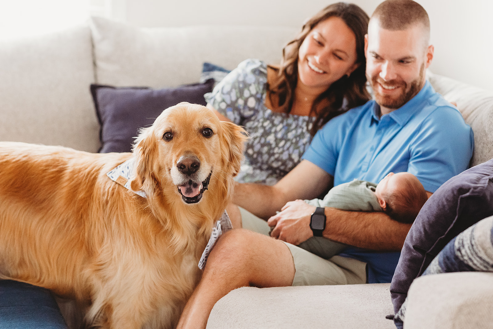 Happy new parents sit on a couch with their sleeping newborn and golden retriever after using RMH Family Birthplace