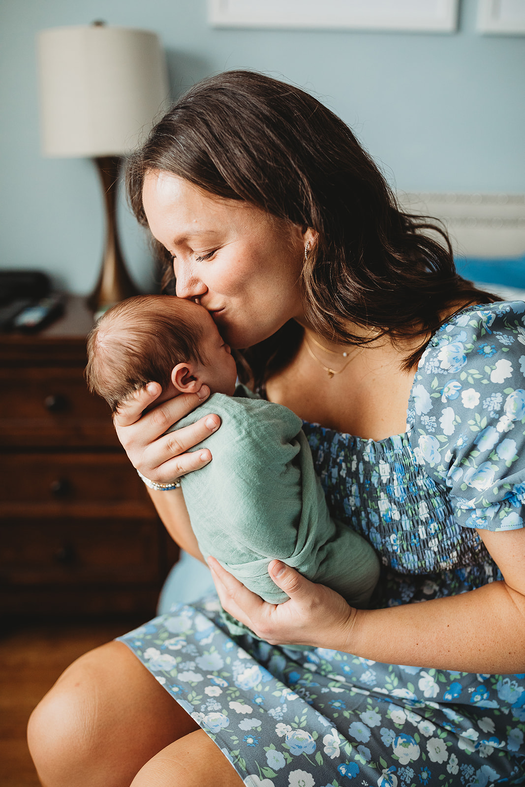 A happy new mom in a blue dress sits on a bed while kissing her sleeping newborn baby after using RMH Family Birthplace