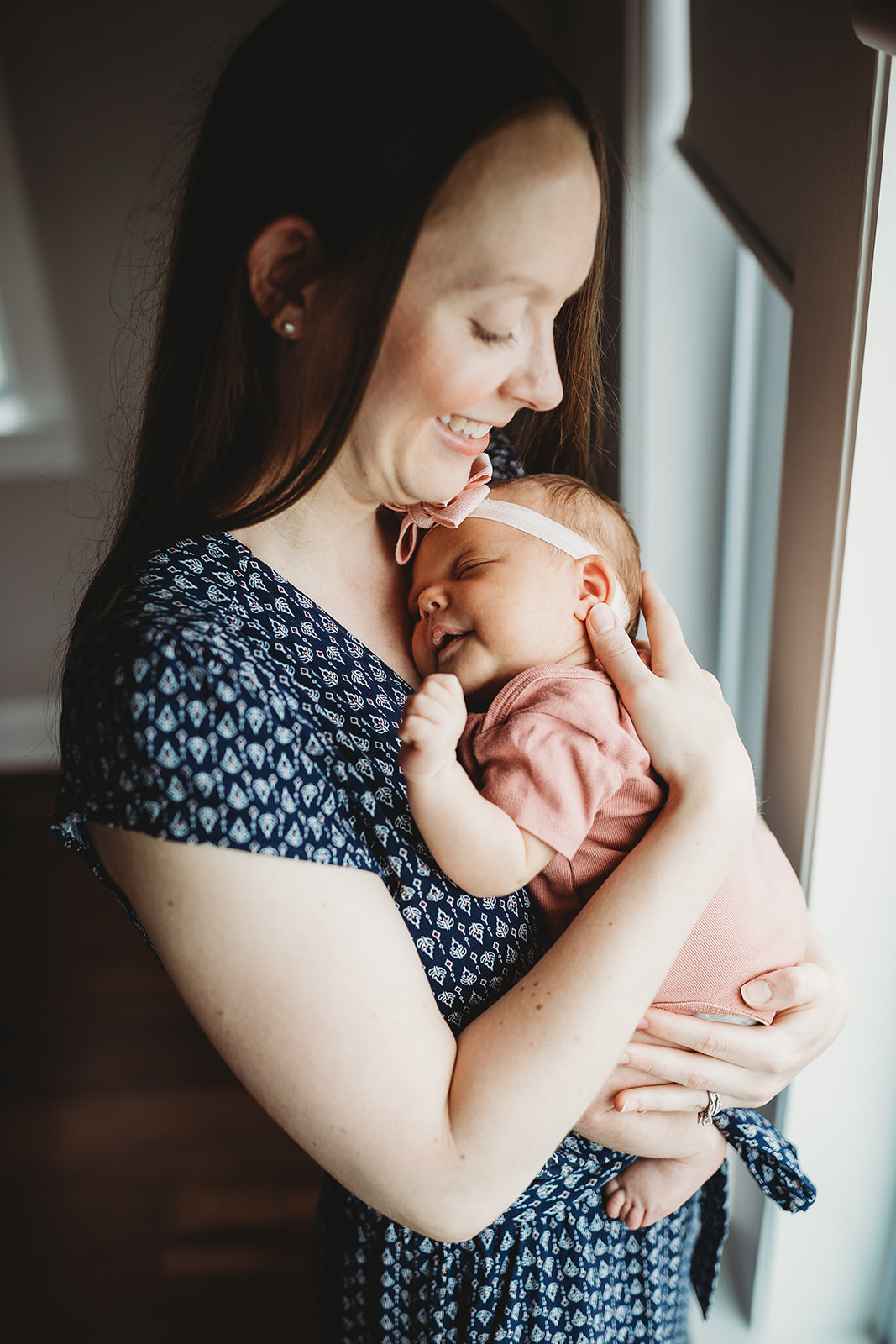 A happy new mom cradles her sleeping newborn daughter against her chest while standing in a window after using Shenandoah Women's Healthcare