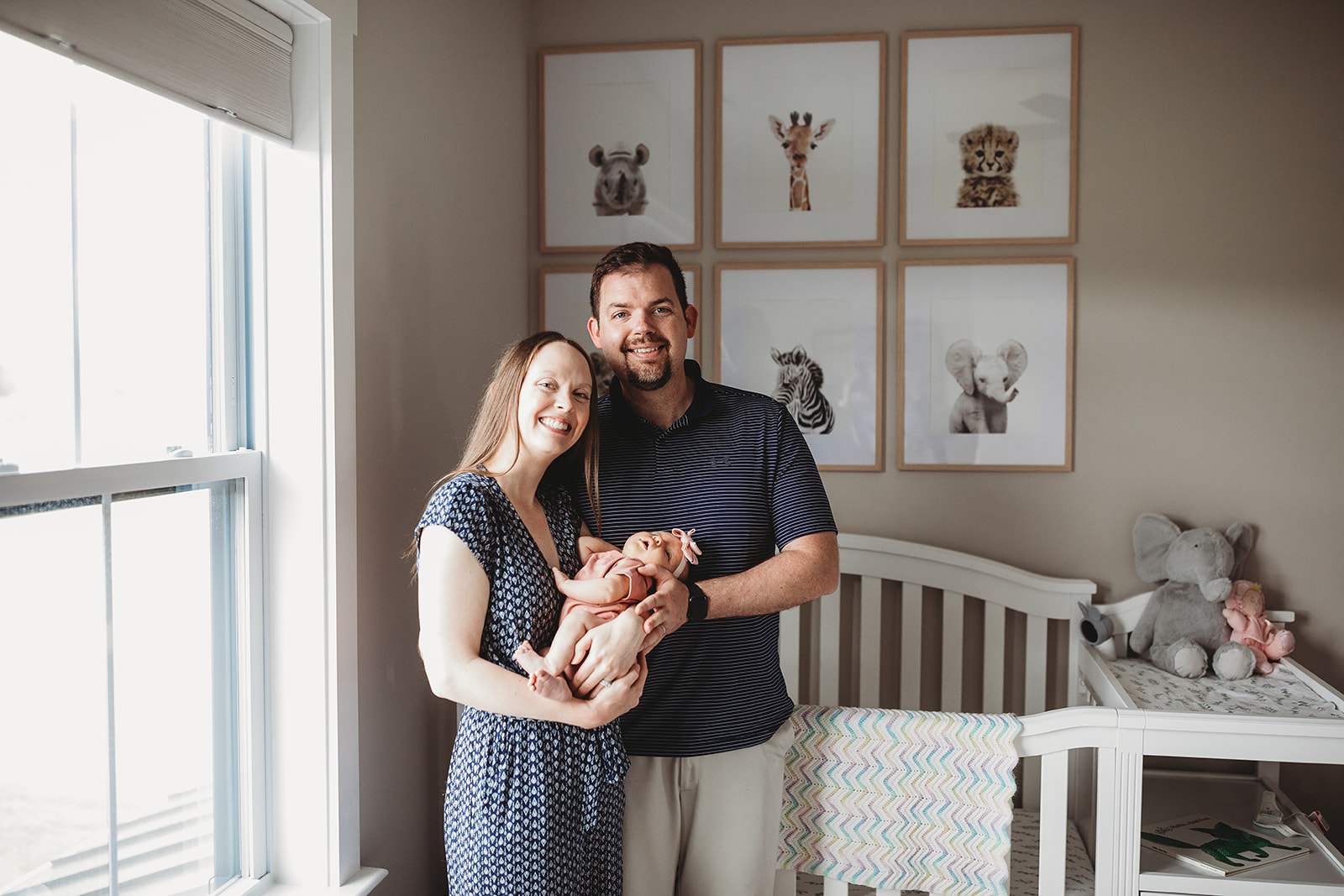 Smiling new parents stand in a nursery with their sleeping newborn baby in their hands