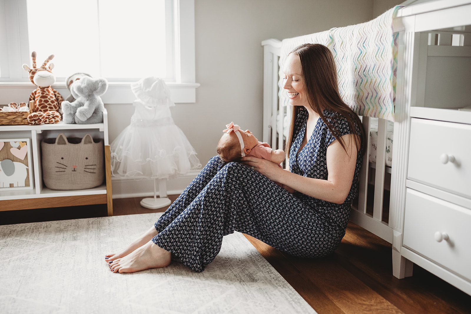 A happy new mom sits against a crib in a nursery with her newborn daughter in her lap after visiting Shenandoah Women's Healthcare