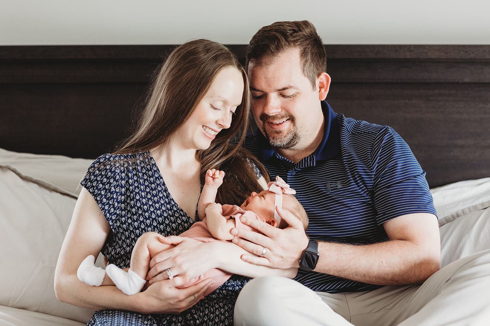 Smiling new parents sit on a bed with their newborn daughter between them