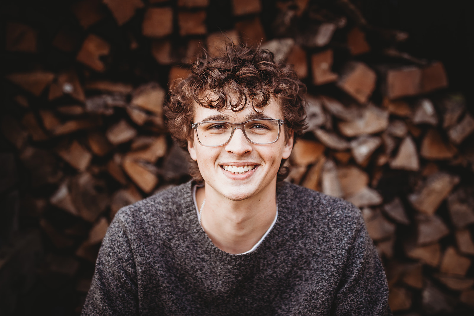 A happy high school senior smiles while sitting by a large stack of firewood