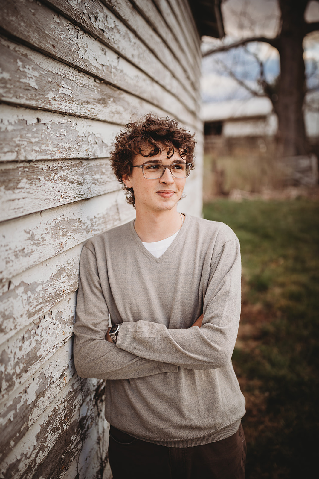 A high school senior leans on a rustic building in a grey long sleeve shirt and smiling over their shoulder